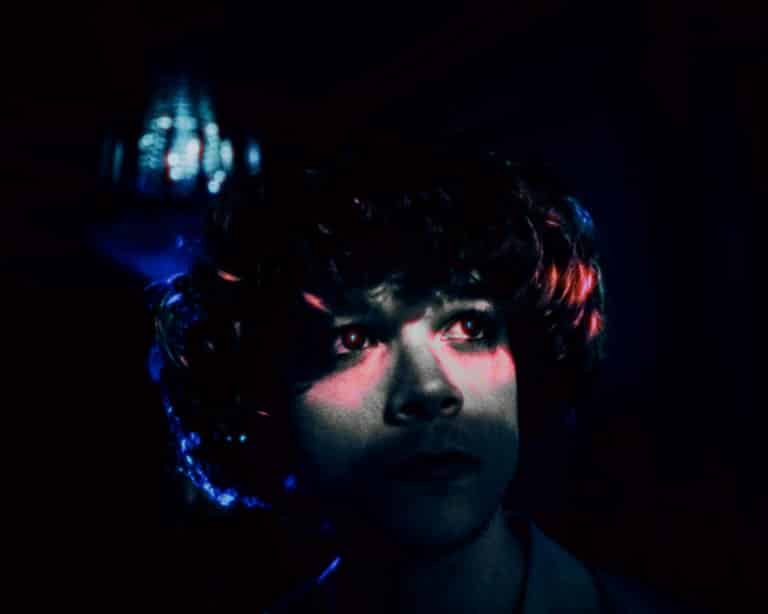 A picture of Indigo Dias of Dispirited Spirits facing sideways on a dark nightly background, a blurry chandelier in the distance, face and eyes partially lit with red highlights, blue shiny silhouette